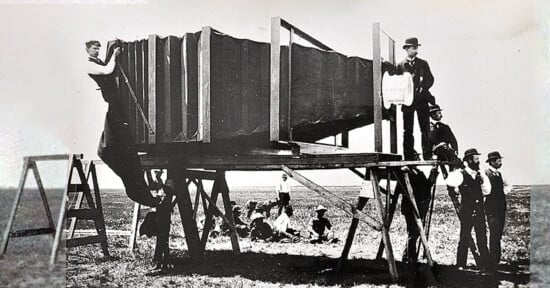 A black and white historical photograph shows several men standing around and on a giant camera, mounted on a large wooden platform. Some men are posing on ladders and support beams, while others sit on the ground in the background.