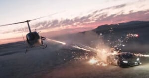 A helicopter fires at a speeding sports car on a desert terrain during sunset, causing sparks and explosions around the vehicle. The scene is intense, with dramatic lighting and a backdrop of mountains and a cloudy sky.