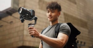 A person with short, curly hair wearing a gray t-shirt smiles while holding a professional camera mounted on a stabilizer. The person is outdoors, with a stone building in the blurred background, and has a black backpack slung over their shoulder.