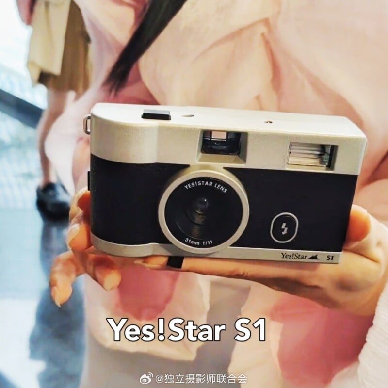 A person, wearing pink, holds a Yes!Star S1 camera with a 31mm f/11 lens. The camera is silver and black with an inbuilt flash, and the text "Yes!Star S1" is prominently displayed below the lens. The brand logo and model are also visible on the camera's face.