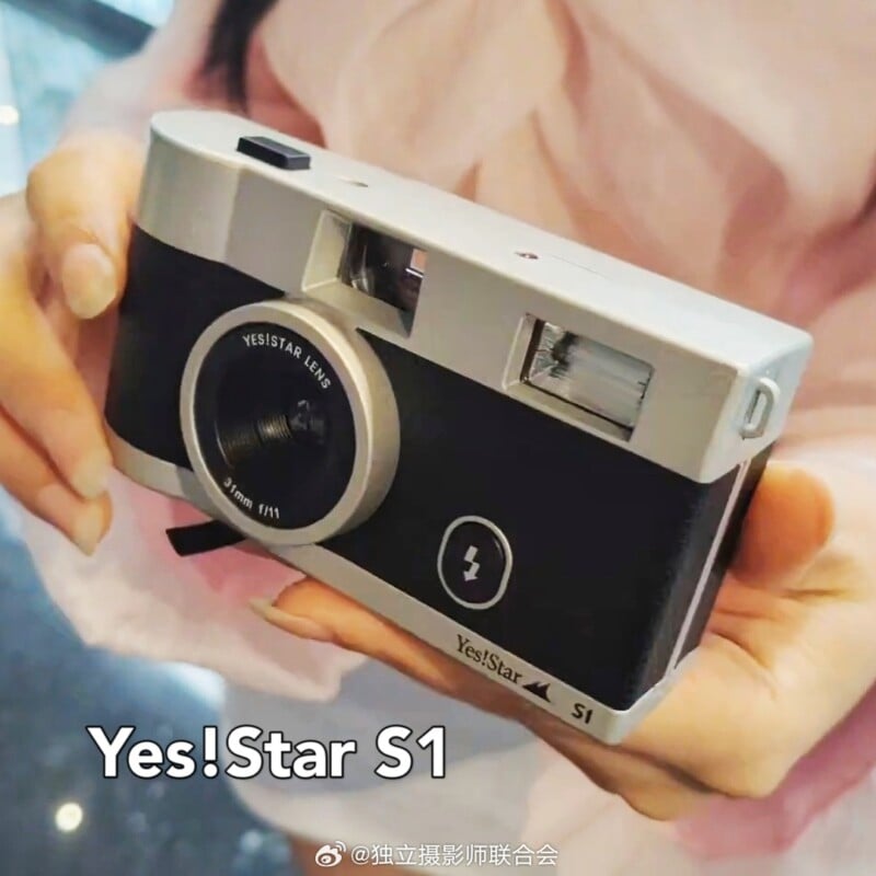 A person holds a vintage-style Yes!Star S1 film camera with both hands. The camera is silver and black with a Yes!Star lens. The background is blurred, and only the person's hands and part of their pink clothing is visible. "Yes!Star S1" text is overlaid on the image.
