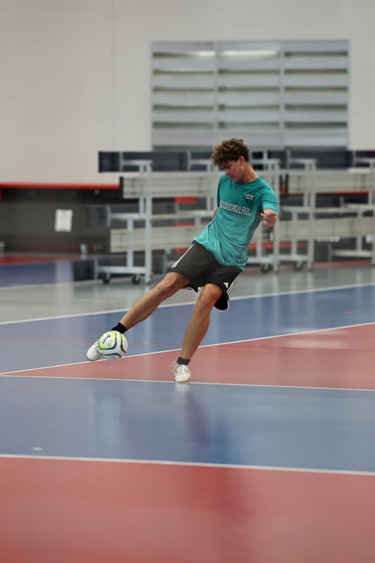 A young man in a teal shirt and black shorts plays indoor soccer on a polished gym floor. He is captured mid-kick, with his right leg extended and his left foot on the ground. The blurred background includes shelves and gymnasium equipment.