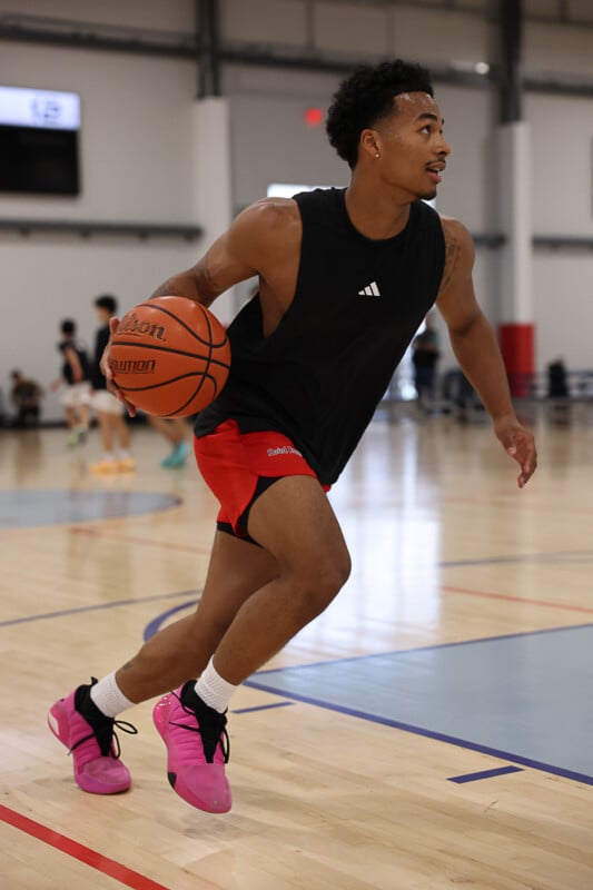 A basketball player in a black sleeveless shirt and red shorts dribbles a ball on an indoor court. He is wearing bright pink sneakers and is looking up as he prepares to make a move. There are other players blurred in the background.