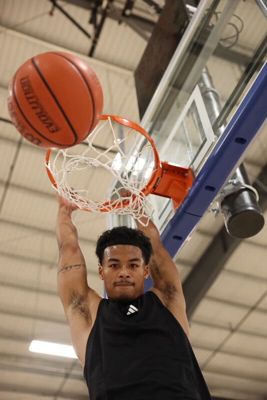 A basketball player in a black tank top jumps and dunks the ball into the hoop at an indoor court. The player's arms are extended, grasping the rim, and their expression is focused. The basketball net and part of the backboard are visible above.