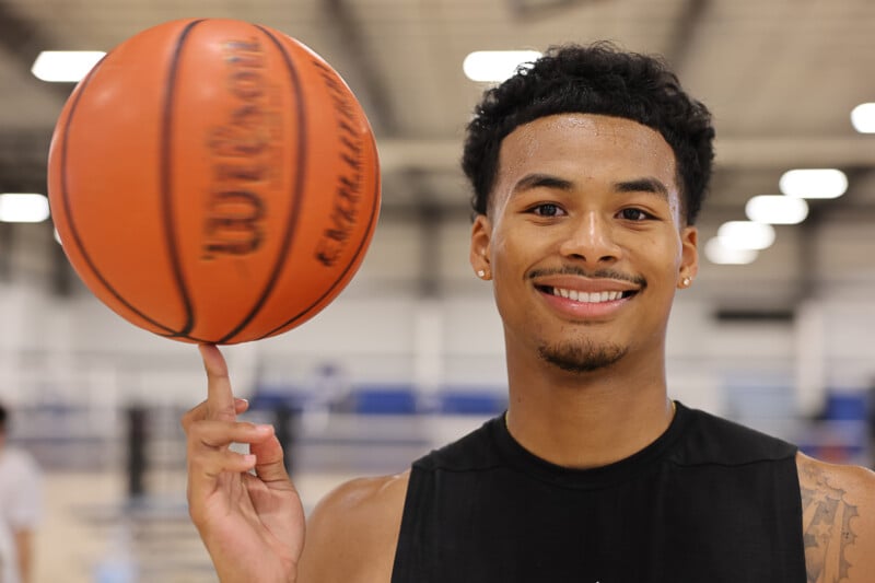 A young man with short black hair and a black sleeveless shirt smiles while spinning a basketball on his finger. He is standing in an indoor sports facility with blurred background elements including a court and gym equipment.