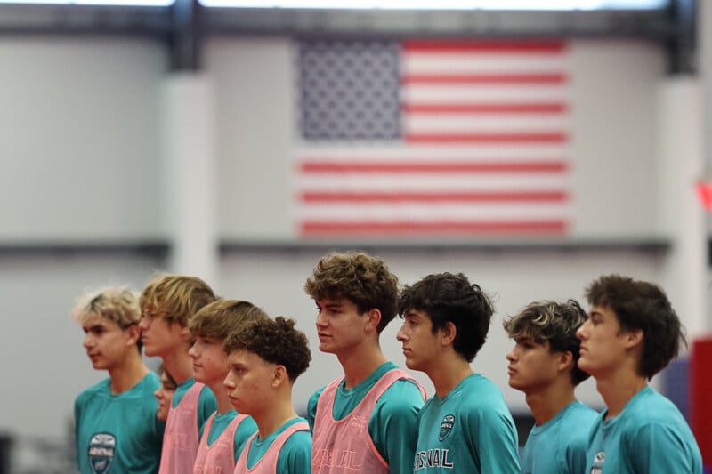 A group of young men wearing teal sports jerseys and pink bibs stand in a line with serious expressions. An American flag is visible in the background, indicating an indoor setting such as a gymnasium.