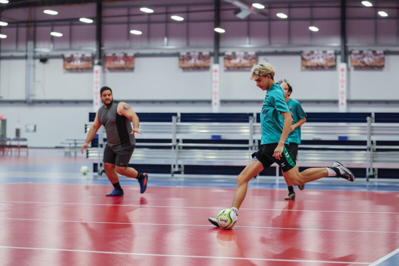 Two people are playing indoor soccer on a red and blue court. One person in a teal shirt is mid-kick towards a soccer ball, while another in a dark outfit is observing in the background. Benches line the wall behind them under a high ceiling with multiple lights.