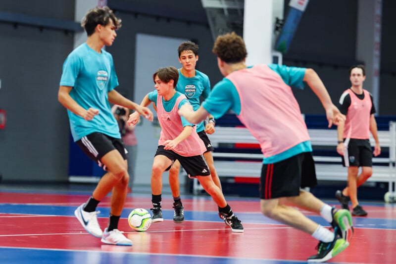 A group of young men play futsal indoors on a blue and red court. One player in a blue jersey tries to maneuver past opponents while controlling the ball. Three others in pink bibs and one in a black jersey are actively participating in the game.