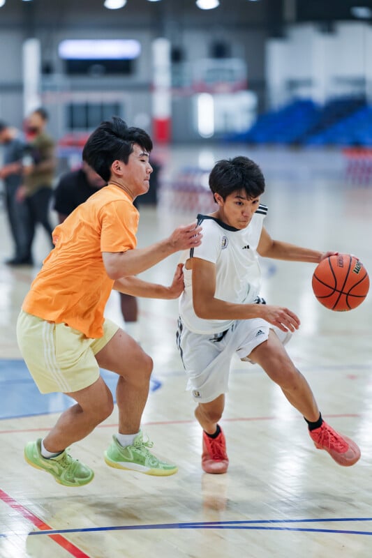 Two young men are playing basketball in an indoor court. The player in orange is attempting to block the player in white, who is dribbling the basketball and driving towards the basket. The background shows an empty area with some spectators watching the game.