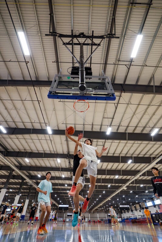 A basketball game is in action inside a large indoor court. A player in a white jersey is leaping toward the hoop for a layup while other players observe or prepare to rebound. The ceiling has bright lights illuminating the game.