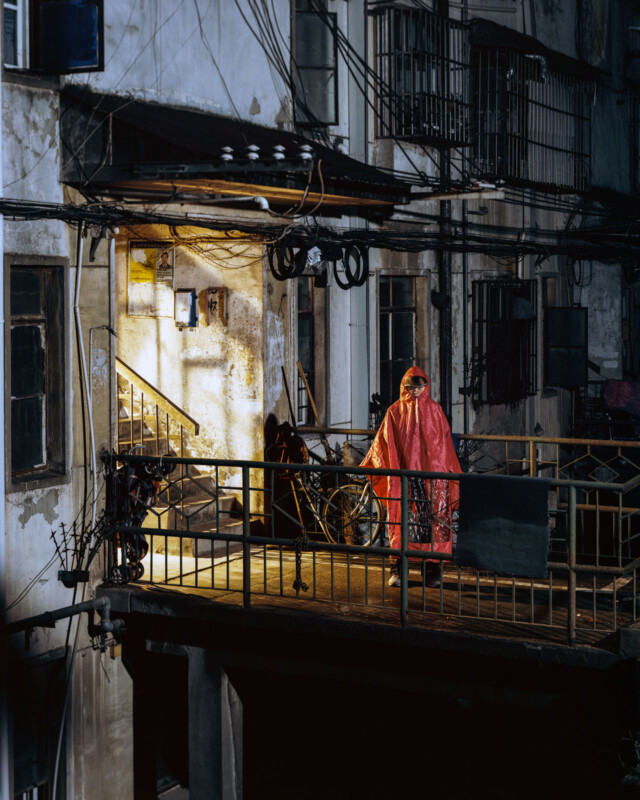 A person wearing a pink raincoat and hat stands on a dimly lit balcony of an old, weathered building at night. The area is cluttered with exposed wiring, bicycles, and surrounding balconies. A warm light emanates from an open doorway inside the building.