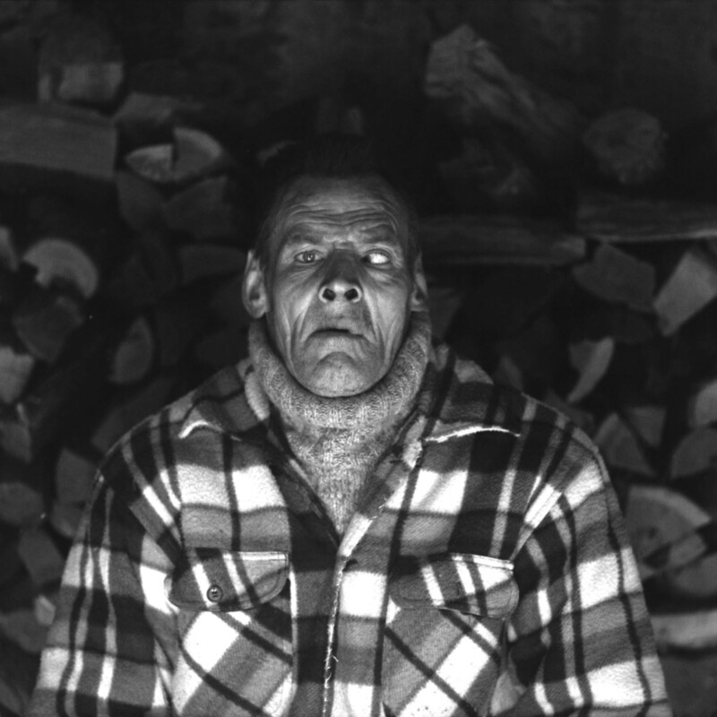 A person with a solemn expression is wearing a plaid shirt and turtleneck sweater, standing in front of a stack of chopped firewood. The background is dark and composed of various logs. The image is in black and white.