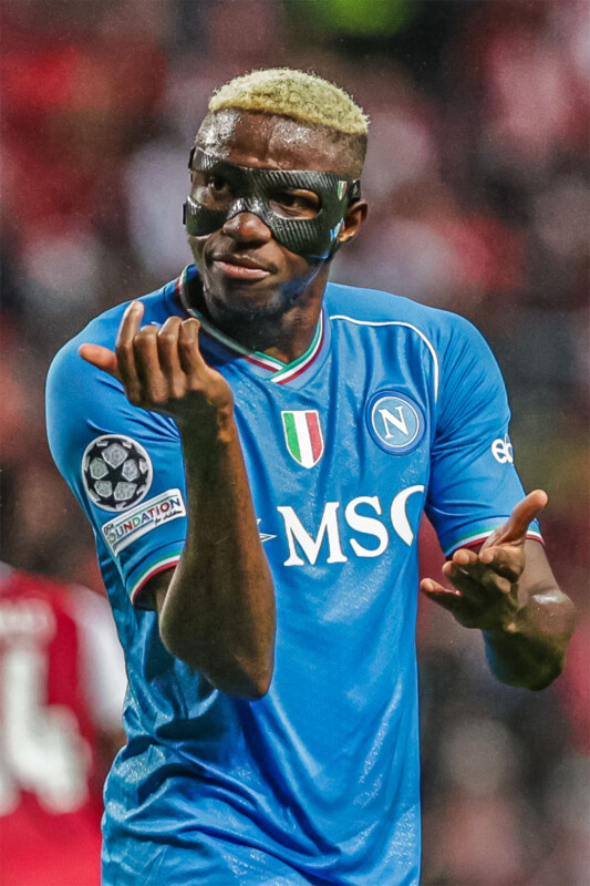 A soccer player wearing a blue Napoli jersey and a protective face mask gestures with his hands on the field. He has short blond hair and is wearing a Champions League patch on his right sleeve. The background shows a blurred crowd of spectators.