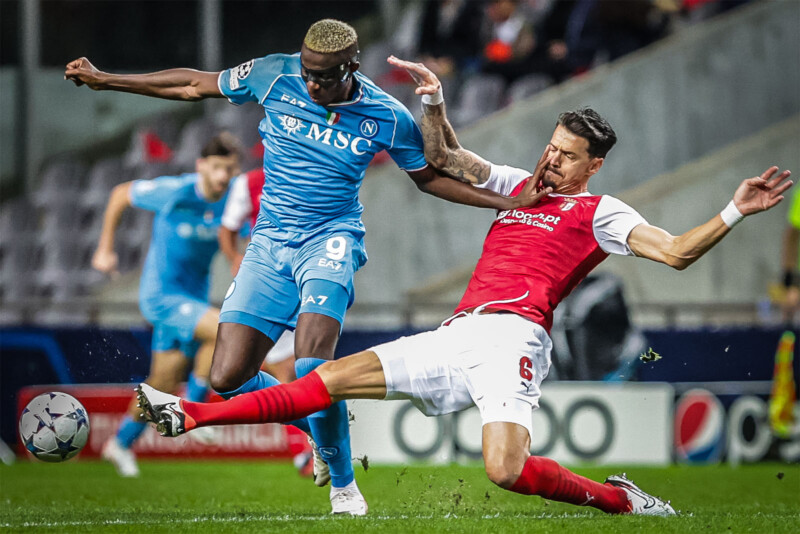 A soccer player in a blue jersey attempts to kick the ball while another player in a red and white jersey slides in attempting to tackle. Both players are intensely focused, and the action occurs on a grassy field with some spectators and lights in the background.