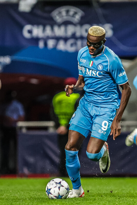 A soccer player in a blue uniform with the number 9 is controlling a ball on the field. He is wearing a face mask, and behind him, blurred, are a sideline official in yellow and a blue "Champions" banner.