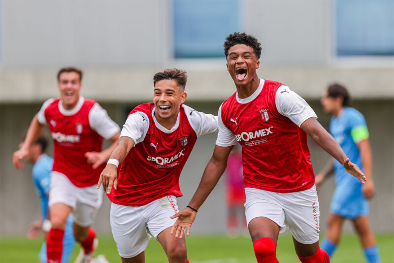 Two soccer players wearing red and white jerseys celebrate a goal, running with arms outstretched and expressions of joy. A teammate follows behind, and opponents in blue jerseys can be seen in the background on the field.