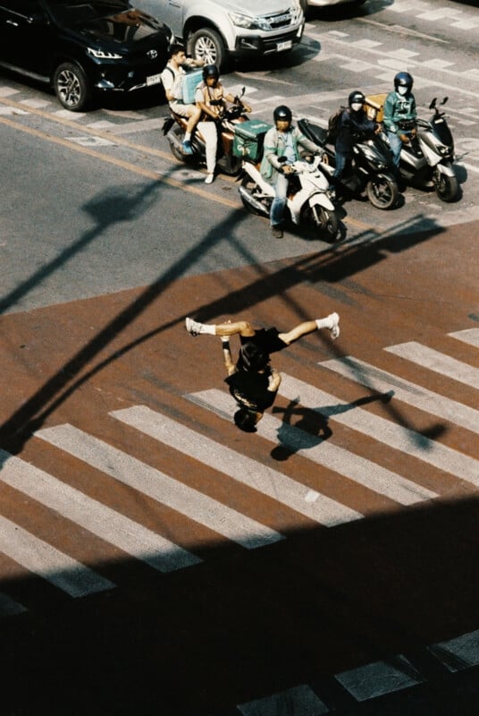 An individual is performing a handstand in the middle of a crosswalk. In the background, four individuals on scooters are waiting at a red light on the road. The shadows cast by nearby structures are visible, creating intersecting lines on the pavement.