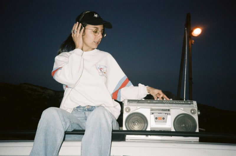 A person wearing a white long-sleeve shirt, blue jeans, glasses, and a black cap sits on a surface while listening to music on a boombox. They hold one hand to their ear and adjust buttons on the boombox with the other hand, with a lamppost in the background at dusk.