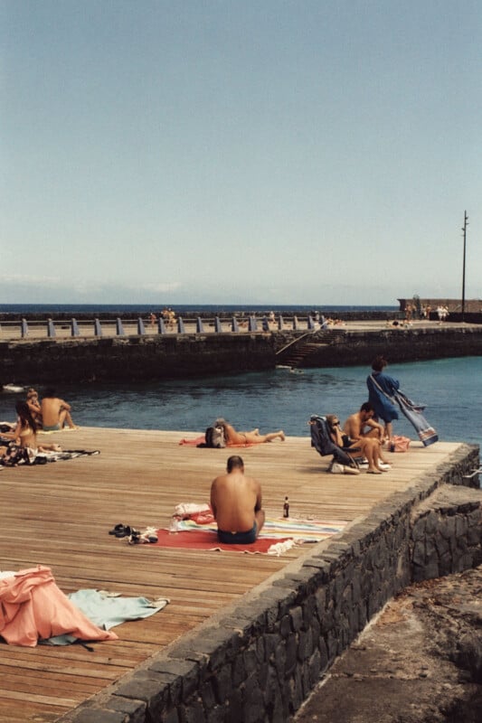 A group of people sunbathing and relaxing on a wooden pier by the water. Towels and chairs are scattered around, and some people are lying down while others are sitting. The pier is surrounded by a low stone wall, and the sky is clear and blue.