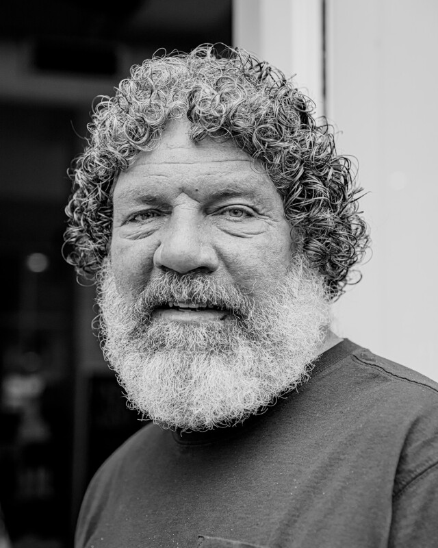 Black and white portrait of a man with curly hair and a full beard. He is wearing a dark-colored shirt and is smiling slightly, looking directly at the camera. The background is blurred, focusing attention on his face.