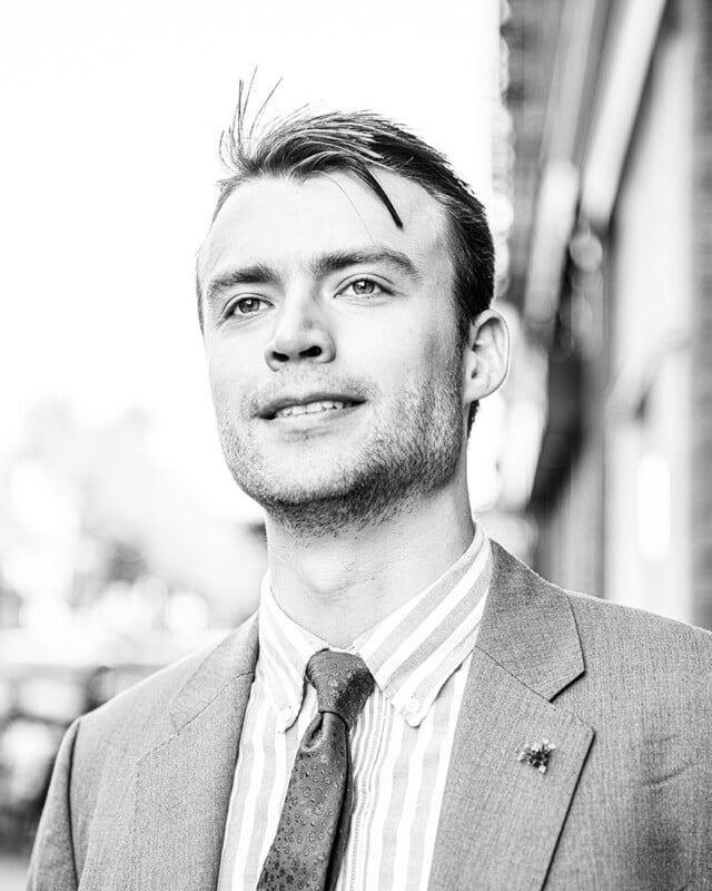 Black and white portrait of a young man wearing a suit, tie, and striped shirt. He is looking slightly upward and smiling. The background appears to be an outdoor urban setting with blurred buildings.