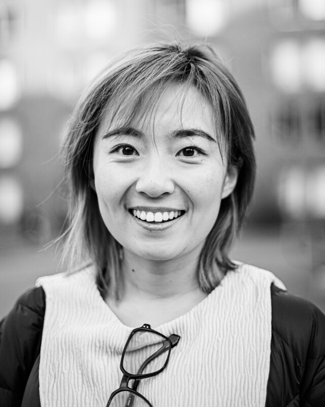 Black and white photo of a person smiling broadly at the camera. They have short, straight hair and are wearing a light-colored, textured top. Glasses are visible hanging from the collar. The background shows blurred buildings.