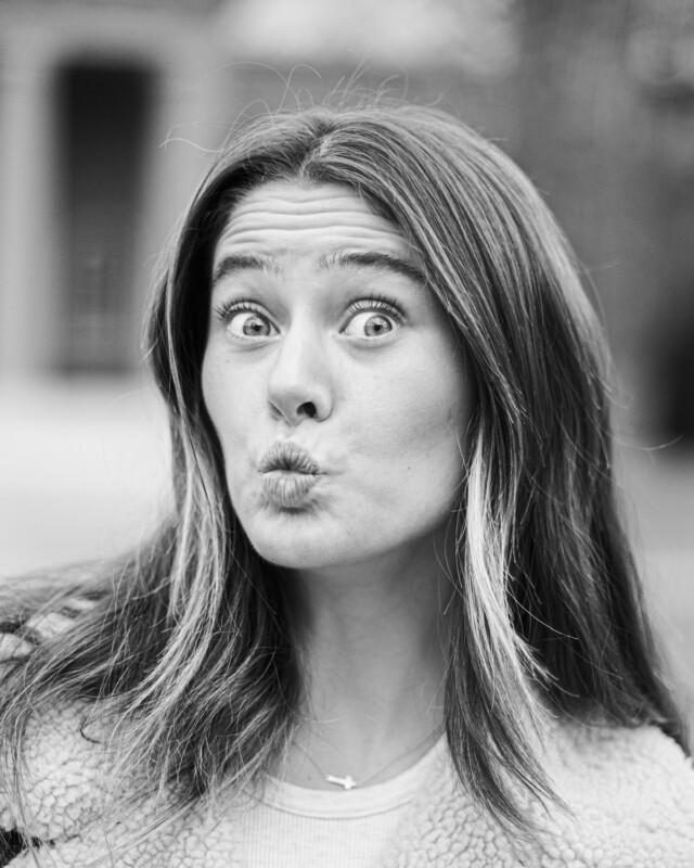 A black and white close-up photograph of a woman with long, straight hair, making a playful, surprised face. Her eyes are wide open, and her lips are puckered as if blowing a kiss. She is wearing a fur-lined jacket and a necklace with a small cross pendant.