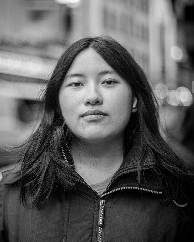 Black and white portrait of a young person with long, dark hair, looking directly at the camera. They are wearing a jacket with a zip-up collar. The background is out of focus, featuring an urban setting with blurred buildings and lights.