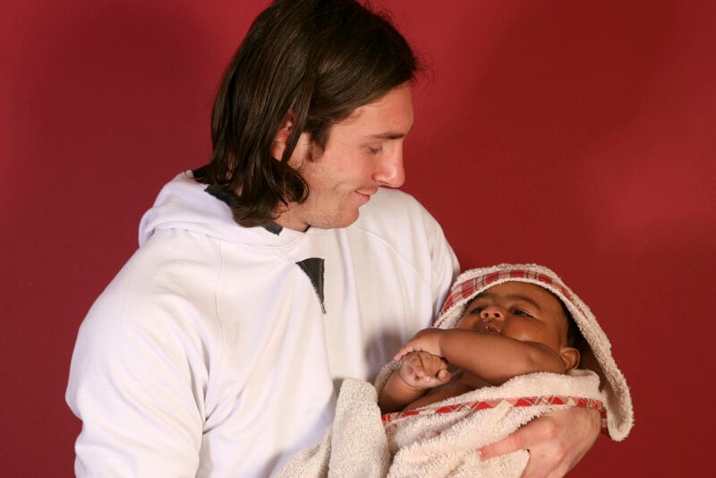 A person with long, dark hair, wearing a white hoodie, is holding a baby wrapped in a beige towel with a red edge. The person is looking affectionately at the baby, who is gazing back. The background is a solid red color.