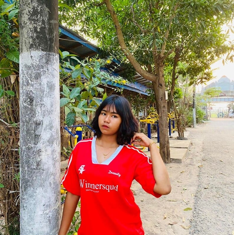 A young woman with shoulder-length black hair stands near a tree trunk, wearing a red shirt with "Winnersquad" printed on it. She holds her hair with her right hand while looking slightly to the left. The background features foliage and a sunlit pathway.