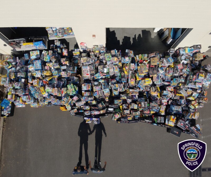 A large collection of assorted toys is spread out in front of a building, casting shadows on the pavement. Two people stand near the toys, and a Springfield Police badge logo is visible in the bottom-right corner of the image.