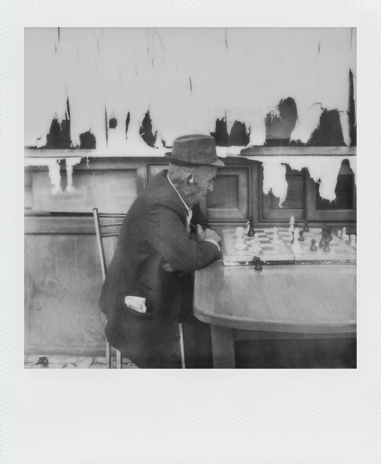 A black-and-white photograph shows an elderly man wearing a hat and a suit sitting at a wooden table. He is intently looking at a chessboard with pieces in play. The background features a wall with significant peeling paint.