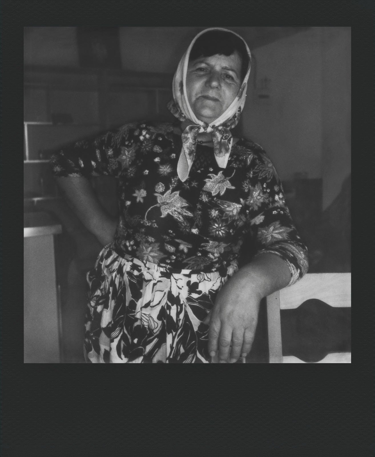 A black and white photo of an older woman leaning on a wooden chair with one hand on her hip. She wears a floral-patterned blouse, a matching skirt, and a headscarf. Her expression is stoic, and she stands in a room with blurry, indeterminate furnishings in the background.