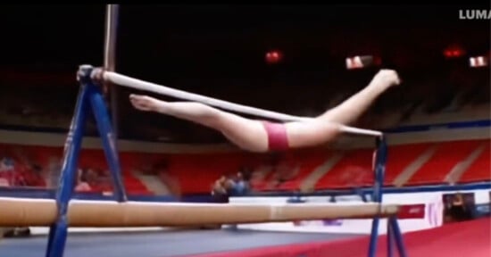 A gymnast in red performing a routine on uneven parallel bars. She is suspended horizontally, with her legs extended out on one bar and her hands gripping the other. The background shows an indoor gymnasium with red seating.
