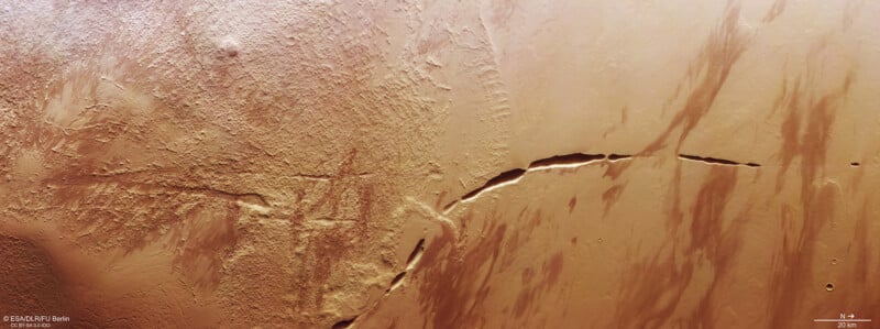 Aerial view of a desert terrain with a large, prominent crack running diagonally from the center towards the bottom right. The surface shows various shades of brown with scattered textures and patterns resembling erosion marks. A scale indicator is at the bottom right.