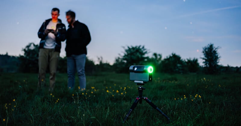 Two people stand in a grassy field at twilight, with a tripod-mounted camera or projector with a green light in front of them. The background shows trees and a darkening sky with a few stars visible.