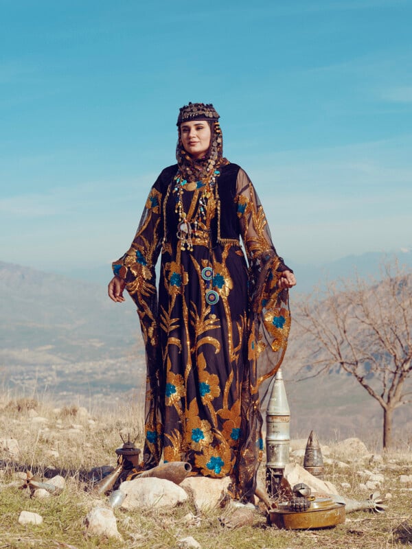 A woman stands outdoors on rocky terrain, wearing a traditional black and gold dress adorned with blue flowers. She has a headscarf and various decorative items around her, with a barren tree and mountains in the background under a clear sky.