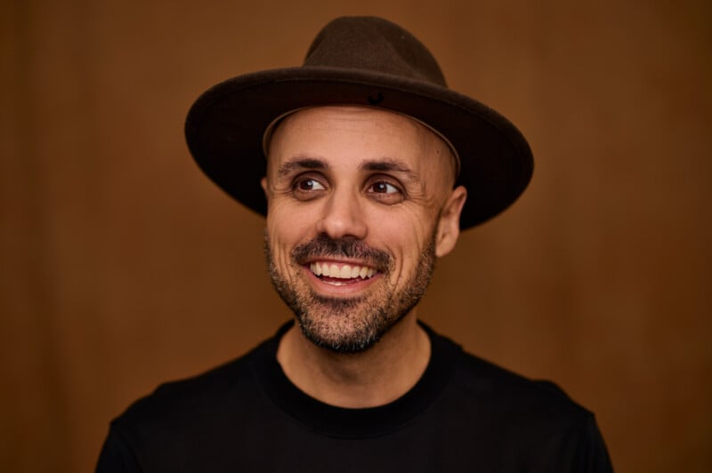 A smiling man with a beard and mustache is wearing a brown hat and a black shirt. The background is a muted brown, providing a warm and neutral backdrop for the subject.