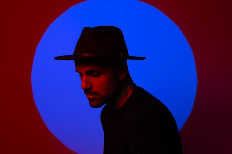 A person in a dark silhouette wears a black hat, standing against a background with a vibrant blue circle amidst a red backdrop. The image highlights the contrast between the subject and the striking blue and red colors.