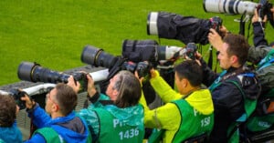 A group of photographers wearing green and blue vests, numbered 1436, 1518, and 158, aim their cameras with large telephoto lenses at an event, possibly a sports game. They are positioned on the sidelines, capturing the action on the green field in front of them.