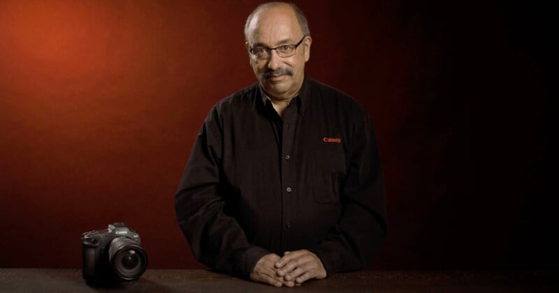 A person with short hair and glasses stands behind a wooden table, wearing a black shirt with "Canon" embroidered on it. They have a relaxed smile and hands resting on the table. A Canon camera is placed on the table to their right against a dark red background.