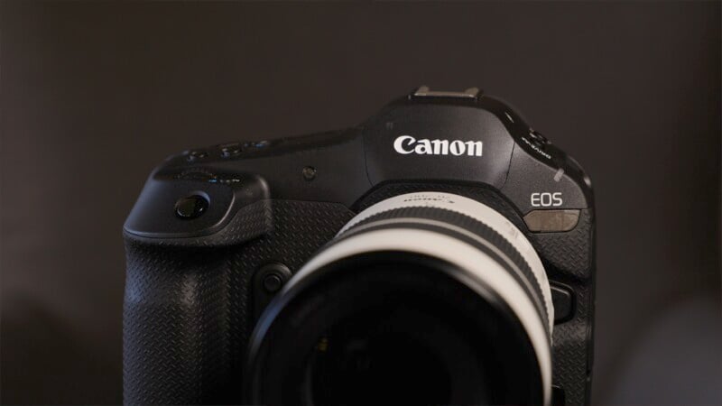 Close-up image of a Canon EOS DSLR camera with a black body and a white lens. The text "Canon" is prominently displayed on the upper front of the camera. The background is a subdued, dark color, highlighting the camera.