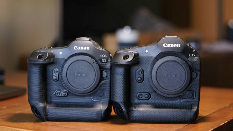 Two Canon DSLR cameras are placed side by side on a wooden surface. Both are black, featuring robust grips and professional designs. The camera on the left is labeled "EOS R3" and the one on the right is labeled "EOS." Both have no lenses attached.