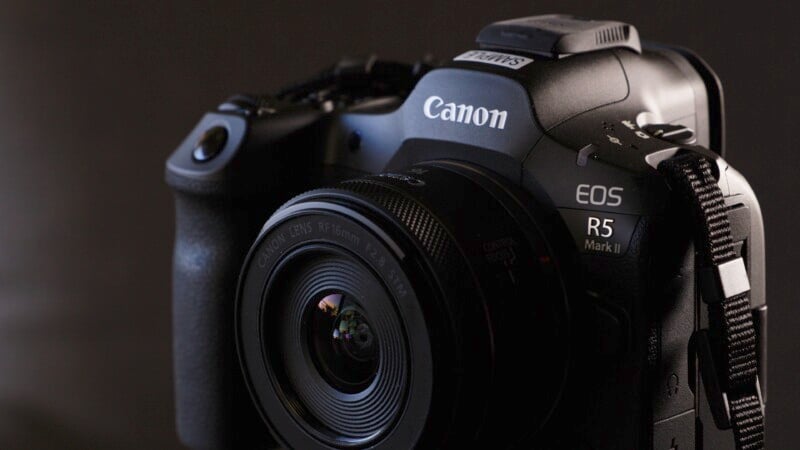 Close-up of a Canon EOS R5 Mark II camera in a dark setting, showcasing its lens, brand logo, and various buttons and features on the camera body. The detailed texture of the grip and the sleek design are highlighted.