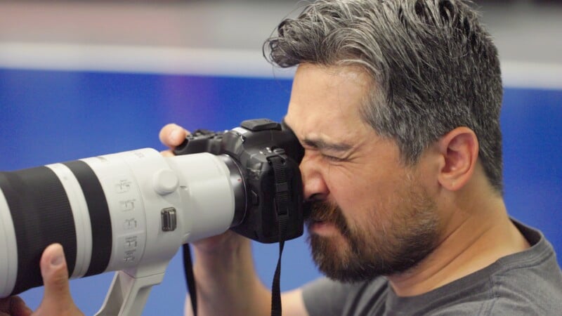 A man with gray and black hair, wearing a gray shirt, is looking through the viewfinder of a large, professional camera with a long telephoto lens. His right eye is closed while he focuses with his left eye. The background is blurry, with shades of blue.