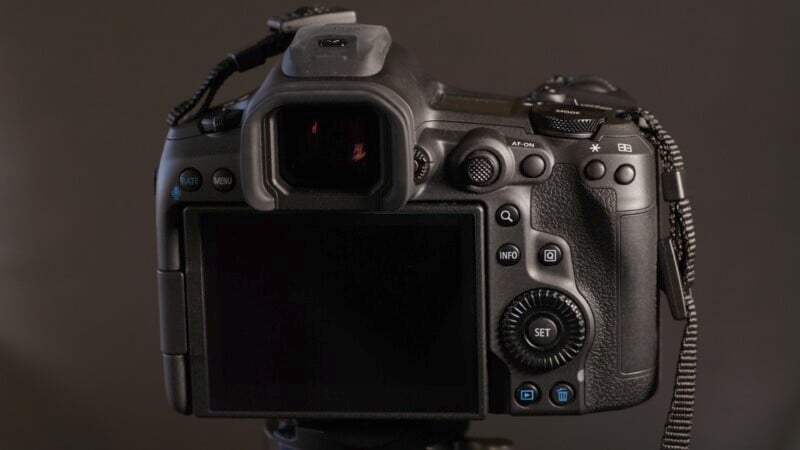 A DSLR camera viewed from the back, showing a large LCD screen, various buttons, dials, and an electronic viewfinder. The camera appears to be black and is mounted on a tripod. A black strap is attached to the camera.