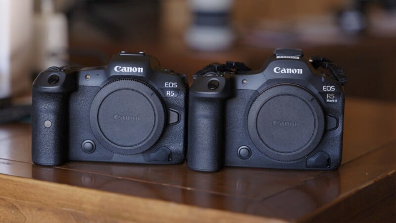 Two Canon cameras, an EOS R5 and an EOS R5 C, are placed side by side on a wooden surface. Both cameras have lens caps on, showing their respective model names on the top right. The background is slightly blurred, emphasizing the cameras.