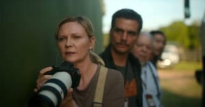 A woman holding a camera with a large lens stands at the forefront, with four people lined up behind her. The group appears focused and intent, possibly observing or waiting for something. The background is outdoors with blurred greenery and sunlight.