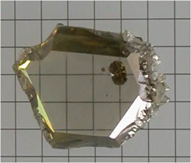 A roughly hexagonal, transparent yellow crystal with jagged edges is displayed against a background of grid paper. The crystal has reflective surfaces and appears angular and slightly irregular in shape.