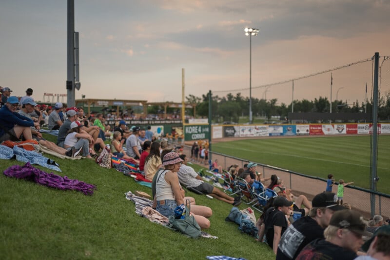 A crowd of people sits on a grassy hill overlooking a baseball field. Some are on blankets and others in lawn chairs, enjoying the warm evening. The field is surrounded by advertisements on the fence, and the sky is a mix of soft clouds and the fading light of sunset.